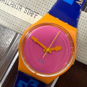 Puede incluir: Un reloj de pulsera Swatch con caja de color naranja brillante y esfera rosa vibrante. El reloj tiene manecillas de hora y minuto naranjas con un segundero amarillo fino. La correa azul muestra un patrón de hojas y el texto Atlanta.