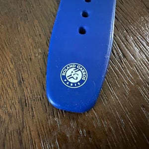 May include: Close-up of a blue watch strap with three holes and a white Roland Garros logo. The strap is made of a flexible material and is set against a wooden surface.