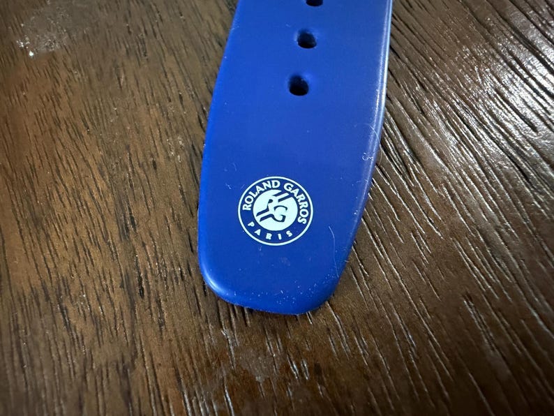 May include: Close-up of a blue watch strap with three holes and a white Roland Garros logo. The strap is made of a flexible material and is set against a wooden surface.