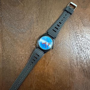 May include: A wristwatch with a black plastic case and a matching black ribbed rubber strap. The watch face features a bright blue background with small yellow star-like accents and three red hands. A silver-toned metal buckle secures the strap.