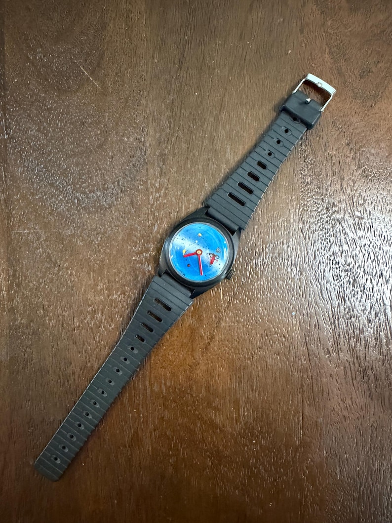 May include: A wristwatch with a black plastic case and a matching black ribbed rubber strap. The watch face features a bright blue background with small yellow star-like accents and three red hands. A silver-toned metal buckle secures the strap.