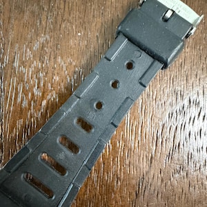May include: A black rubber watch strap featuring a series of rectangular and circular adjustment holes. The strap includes a black rubber keeper loop and a silver-toned metal buckle clasp with the words STAINLESS STEEL engraved on the metal surface.