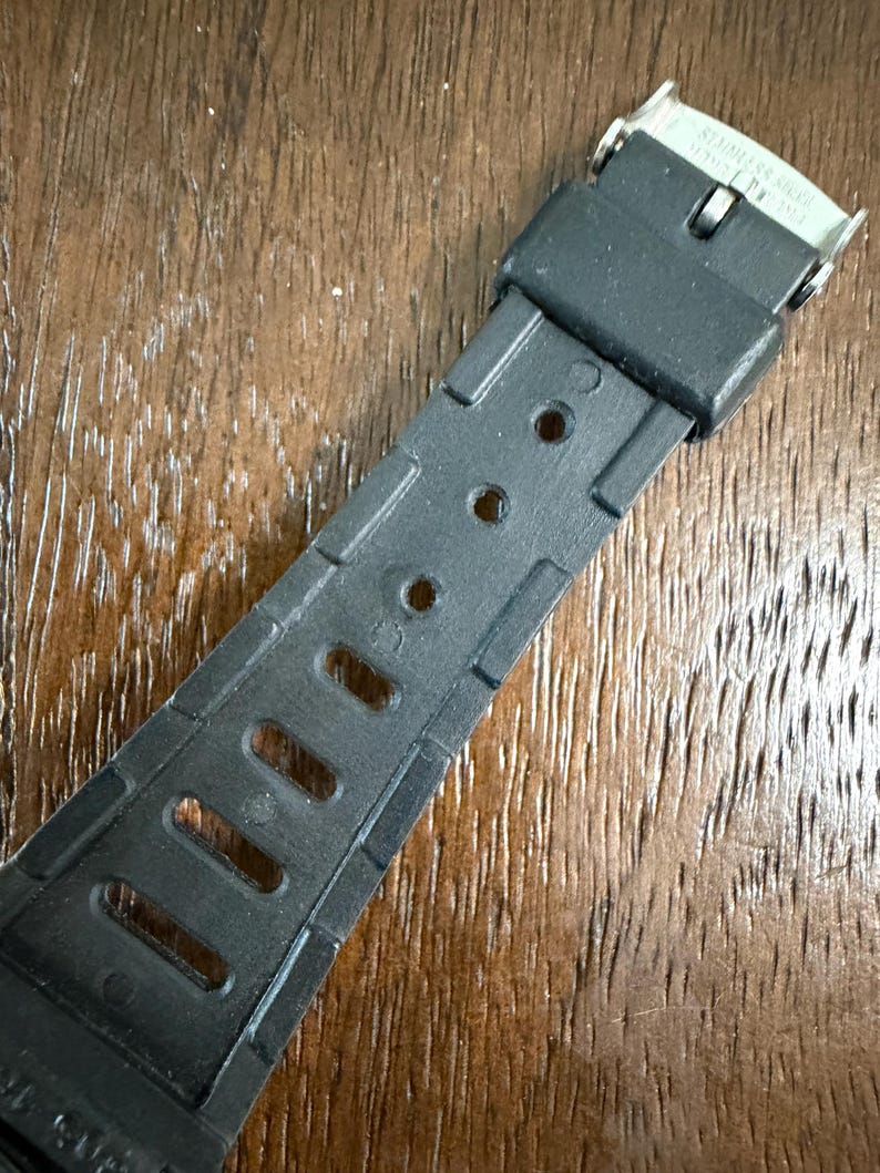 May include: A black rubber watch strap featuring a series of rectangular and circular adjustment holes. The strap includes a black rubber keeper loop and a silver-toned metal buckle clasp with the words STAINLESS STEEL engraved on the metal surface.
