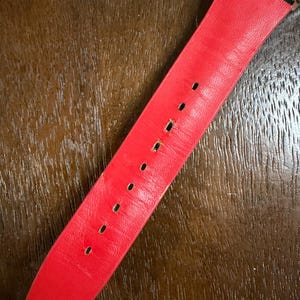 May include: A bright red watch strap featuring a smooth, matte finish with a series of evenly spaced rectangular adjustment holes running down the center. The strap is attached to a metallic watch casing at the top with a dark green connector.