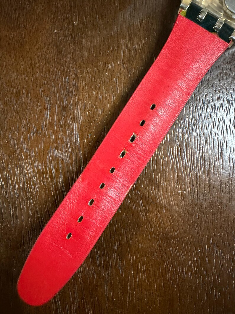 May include: A bright red watch strap featuring a smooth, matte finish with a series of evenly spaced rectangular adjustment holes running down the center. The strap is attached to a metallic watch casing at the top with a dark green connector.