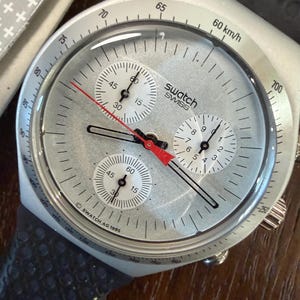 May include: A silver Swatch chronograph wristwatch with a tachymeter scale on the bezel, three white sub-dials, black hour and minute hands, and a red second hand. The dial features the text Swatch Swiss and Swatch AG 1995. It has a dark strap.