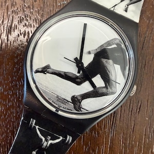 Puede incluir: Un reloj de pulsera Swatch con un estampado gráfico en blanco y negro de atletas en movimiento en la esfera y la correa. La esfera muestra a un corredor, mientras que la correa presenta a un saltador de altura y a un levantador de pesas.
