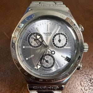 May include: A silver-toned Swatch chronograph wristwatch with a metallic dial, three sub-dials, and a date window at the four o'clock position. The bezel features engraved text, and the watch includes a matching silver-toned metal link bracelet.