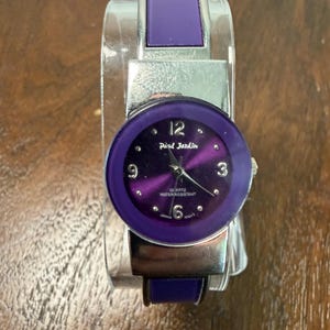 May include: A Paul Jardin quartz watch with a round purple face, silver-tone hands, and numerals at 3, 6, 9, and 12. The watch features a silver-tone metal band with purple inlay accents. The dial is marked with "Paul Jardin", "Quartz", and "Water Resistant".
