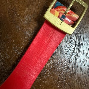 May include: A bright red watch strap featuring a gold-toned metal buckle. The strap is embossed with the word Swatch. The buckle is also engraved with the word Swatch. A portion of the strap near the buckle displays a colorful marbled pattern.