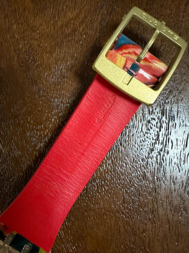 May include: A bright red watch strap featuring a gold-toned metal buckle. The strap is embossed with the word Swatch. The buckle is also engraved with the word Swatch. A portion of the strap near the buckle displays a colorful marbled pattern.