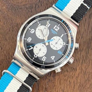 May include: A Swatch chronograph watch with a black dial, white sub-dials, and a blue and white striped nylon strap. The watch face features the number 12, and the word "swatch". The watch has a stainless steel case.