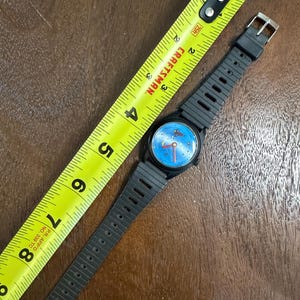 May include: A black wristwatch with a blue dial featuring a small red planet graphic and red hands. The watch has a ribbed black plastic strap and is positioned next to a yellow Craftsman measuring tape showing a length of approximately 20 cm.