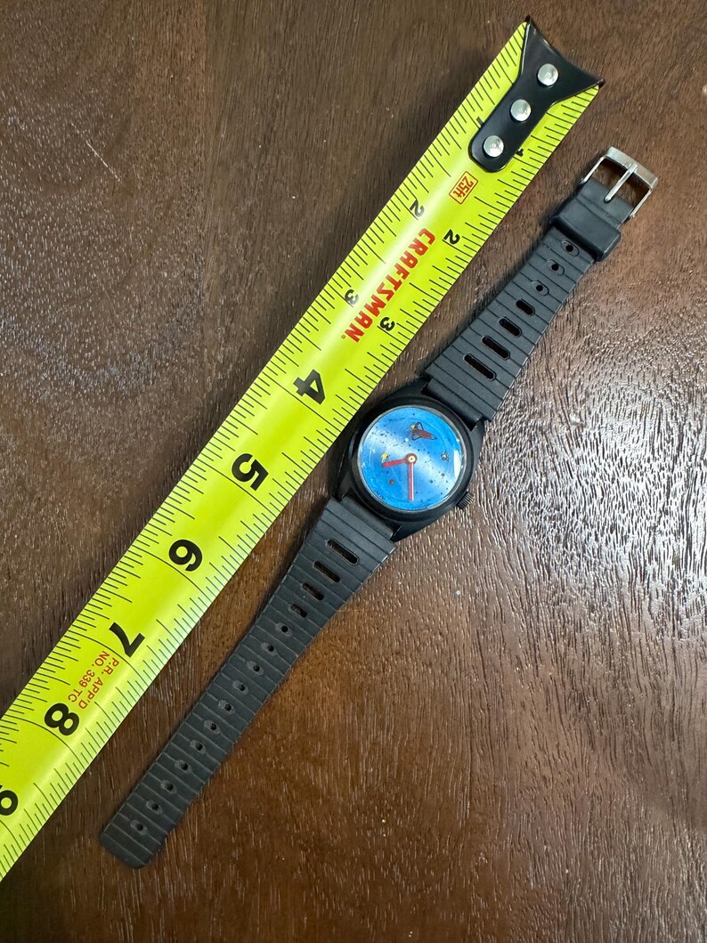 May include: A black wristwatch with a blue dial featuring a small red planet graphic and red hands. The watch has a ribbed black plastic strap and is positioned next to a yellow Craftsman measuring tape showing a length of approximately 20 cm.