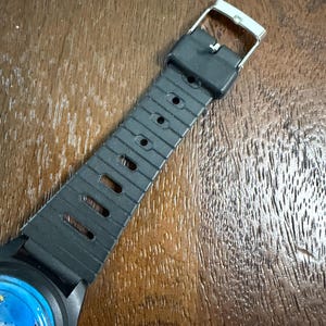 May include: A black rubber watch strap with a series of rectangular and circular adjustment holes, featuring a silver-toned metal buckle. The strap is attached to a watch case that displays a blue dial with a colourful graphic illustration.