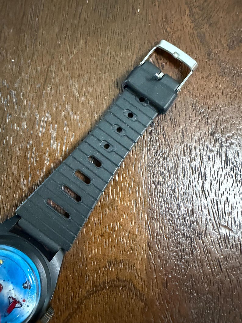May include: A black rubber watch strap with a series of rectangular and circular adjustment holes, featuring a silver-toned metal buckle. The strap is attached to a watch case that displays a blue dial with a colourful graphic illustration.