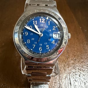 May include: A silver-tone Swatch wristwatch with a blue dial featuring white Arabic numerals for 1 through 12 and smaller 13 through 24 hour markers. The bezel is engraved with the word Swatch, and the watch includes a date window at the 3 o'clock position.
