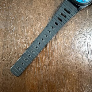 May include: A wristwatch with a black rubber strap featuring a series of rectangular ventilation holes. The watch face is circular with a bright blue background, red hour and minute hands, and small yellow and red graphic details on the dial.