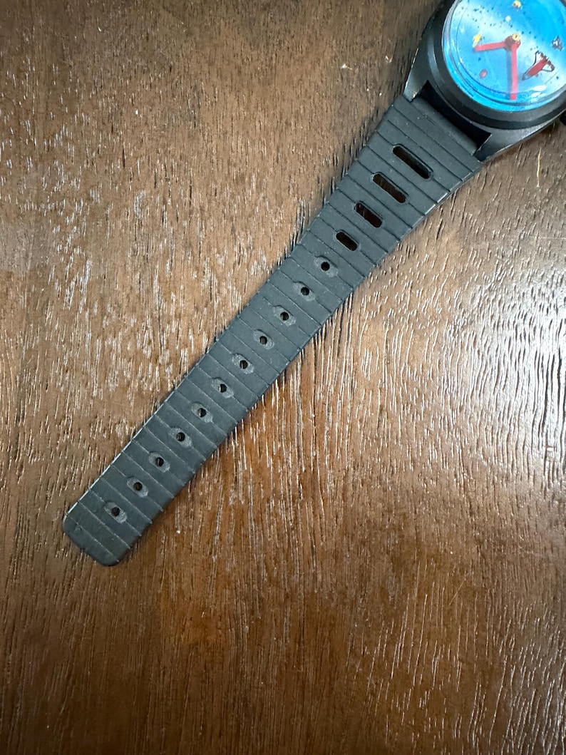 May include: A wristwatch with a black rubber strap featuring a series of rectangular ventilation holes. The watch face is circular with a bright blue background, red hour and minute hands, and small yellow and red graphic details on the dial.