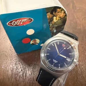 Puede incluir: Un reloj de pulsera Swatch 007 40th Anniversary con esfera azul oscuro, manecillas plateadas y ventana de fecha en la posición de las tres. El bisel plateado presenta nombres de ciudades grabados y la correa es negra con costuras blancas.