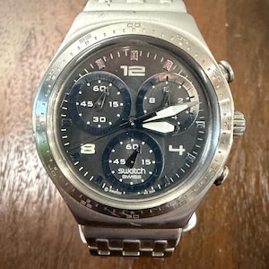 May include: A silver-toned Swatch chronograph watch with a dark grey dial, featuring three sub-dials, luminous hands, and a metallic link bracelet. The watch face displays the number 12 at the top, with the text Swatch Swiss and AG 2004 printed.