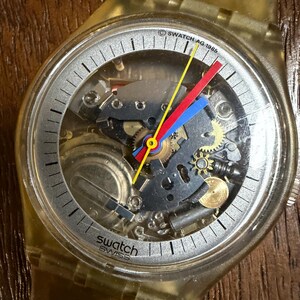 Puede incluir: Un reloj de pulsera Swatch vintage con caja y correa de plástico amarillo translúcido. La esfera esqueleto revela los engranajes internos, con un segundero amarillo, un minutero rojo y una manecilla de hora azul. El texto dice Swatch Swiss.