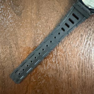 May include: A black watch strap made of a flexible material featuring a series of rectangular and circular adjustment holes. The strap is attached to a metallic watch case back that displays engraved text and circular indentations for a tool.
