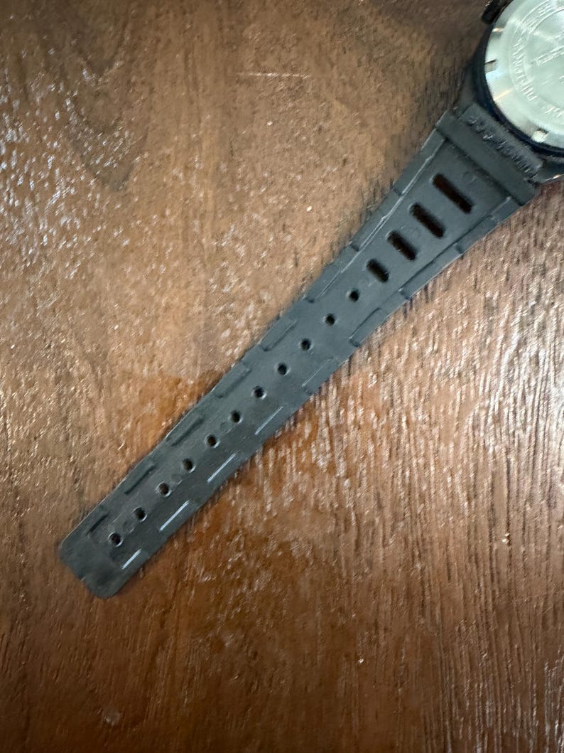 May include: A black watch strap made of a flexible material featuring a series of rectangular and circular adjustment holes. The strap is attached to a metallic watch case back that displays engraved text and circular indentations for a tool.