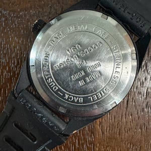 May include: The back of a black wristwatch with a circular stainless steel case back. The text reads: BASE METAL CASE, STAINLESS STEEL BACK, DUSTPROOF, MADE IN HONG KONG FOR GOODS, BOSTON USA. A black rubber strap is attached to the watch casing.