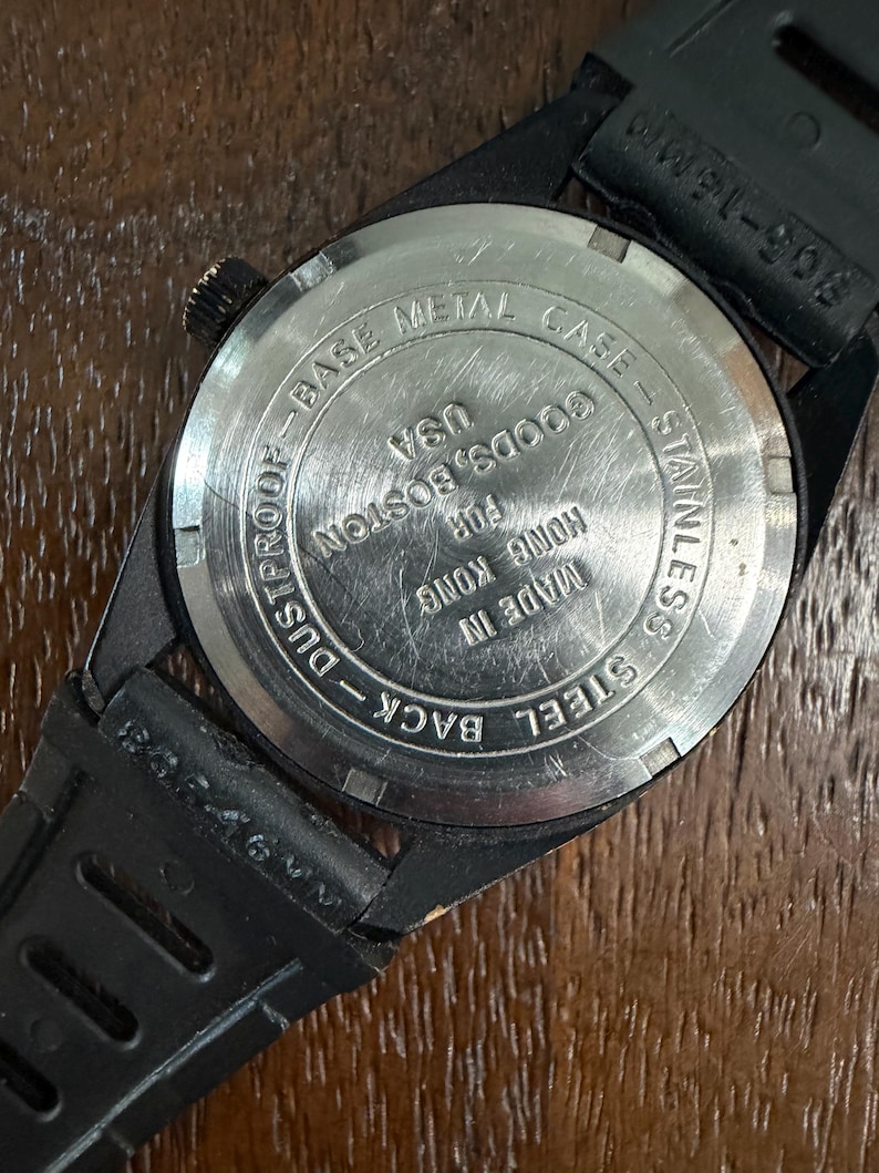 May include: The back of a black wristwatch with a circular stainless steel case back. The text reads: BASE METAL CASE, STAINLESS STEEL BACK, DUSTPROOF, MADE IN HONG KONG FOR GOODS, BOSTON USA. A black rubber strap is attached to the watch casing.