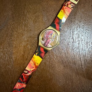 May include: A Swatch wristwatch featuring a gold-tone case and a flexible strap decorated with a vibrant, swirling marbled pattern in shades of red, yellow, and black. The watch face displays a matching abstract design with gold-tone hands.