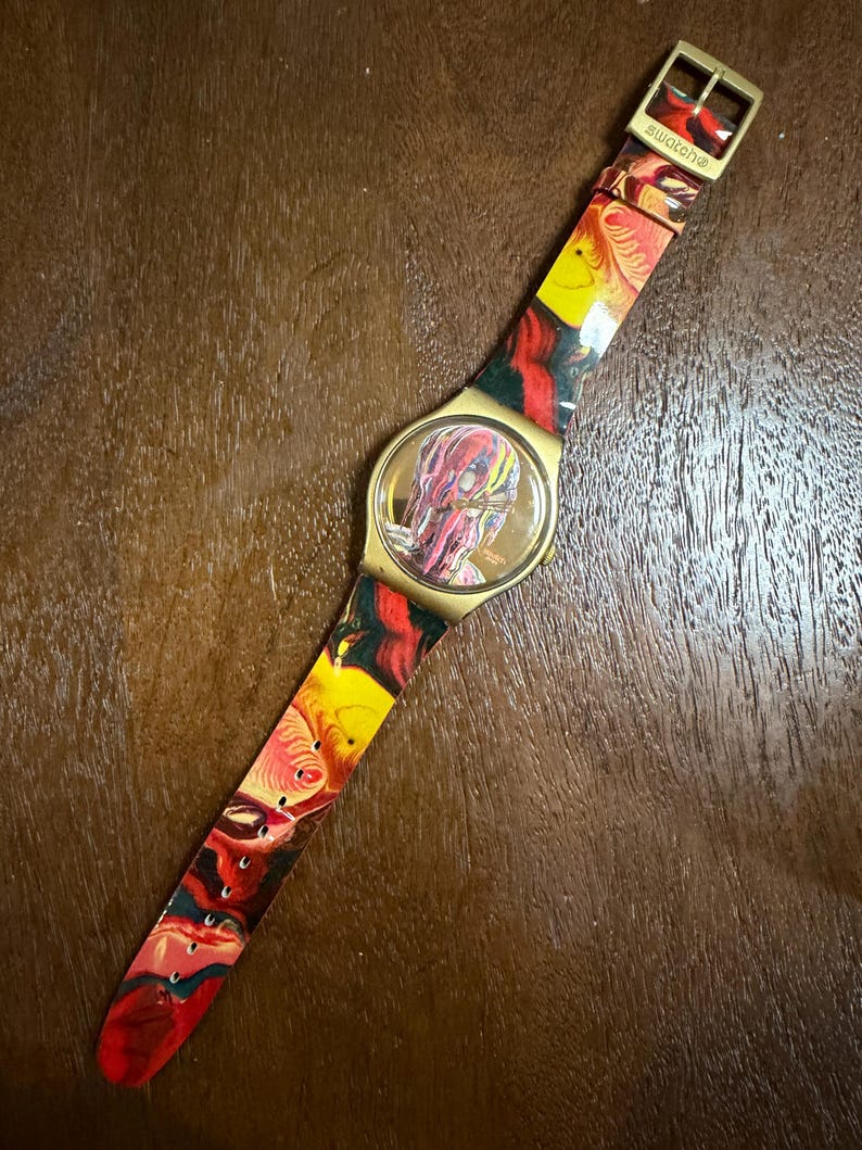 May include: A Swatch wristwatch featuring a gold-tone case and a flexible strap decorated with a vibrant, swirling marbled pattern in shades of red, yellow, and black. The watch face displays a matching abstract design with gold-tone hands.
