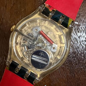 May include: A Swatch wristwatch with a transparent plastic case revealing the internal quartz movement, including a silver battery, a red coil, and metallic gears. The watch features a bright red strap with black and yellow patterned accents.