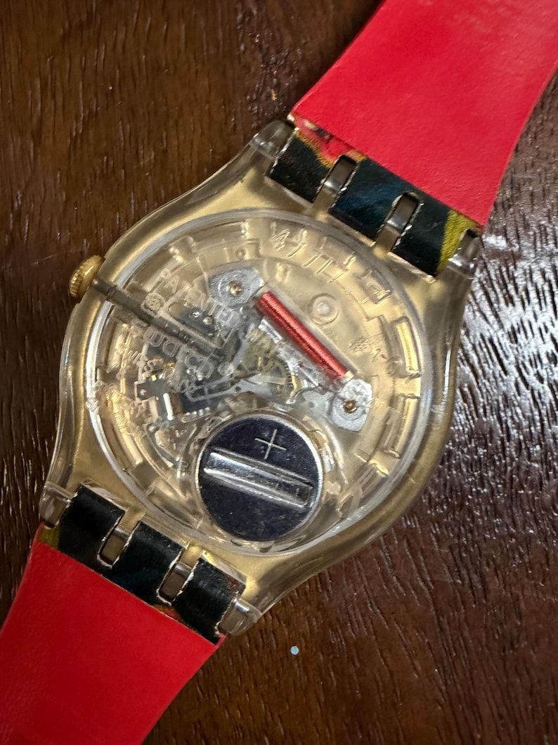 May include: A Swatch wristwatch with a transparent plastic case revealing the internal quartz movement, including a silver battery, a red coil, and metallic gears. The watch features a bright red strap with black and yellow patterned accents.