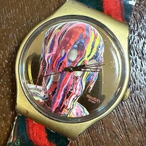 May include: A Swatch wristwatch with a gold-toned case and a clear crystal face. The dial features a graphic of a head covered in dripping, multi-colored paint in shades of red, blue, and yellow. The watch band has a matching abstract paint pattern.