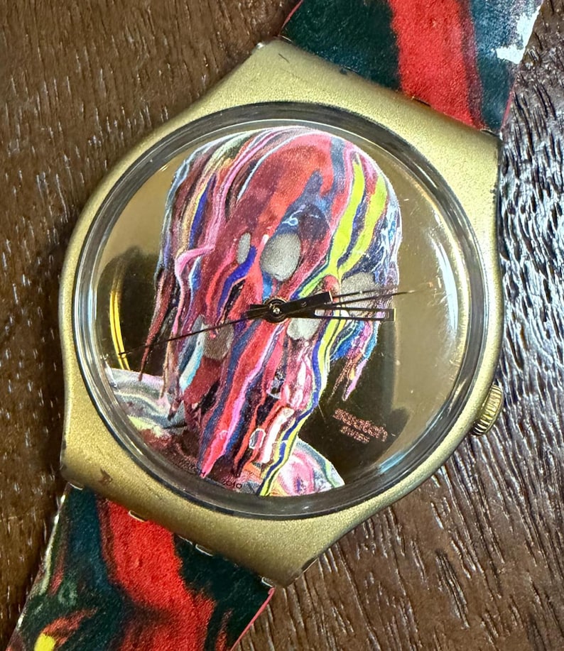 May include: A Swatch wristwatch with a gold-toned case and a clear crystal face. The dial features a graphic of a head covered in dripping, multi-colored paint in shades of red, blue, and yellow. The watch band has a matching abstract paint pattern.