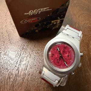 May include: A silver-toned chronograph wristwatch with a vibrant red dial featuring three sub-dials and a metallic band with red accents. The watch is displayed next to its original brown packaging box, which is labeled with the text Tomorrow Never Dies 007.