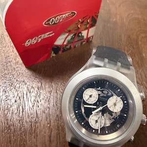 May include: A Swatch chronograph wristwatch featuring a dark blue dial with a world map graphic, three white sub-dials, and a translucent silver case with a black strap, accompanied by a red 007 40th Anniversary box with white text.