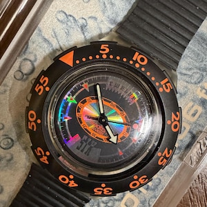 Pode incluir: Um relógio de pulso preto com uma luneta rotativa laranja com marcadores numéricos de 5 a 55. O mostrador exibe um círculo arco-íris holográfico central, ponteiros pretos e o texto Swatch Swiss. Inclui uma pulseira de silicone preta.
