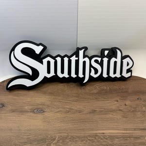 以下が含まれることがあります： 太い黒の縁取りが施された、白のゴシック体フォントで「Southside」という文字が描かれた立体的なウォールサイン。文字は層状になっており、黒い背景の上に白いテキストが浮き上がるような奥行きのあるデザインです。