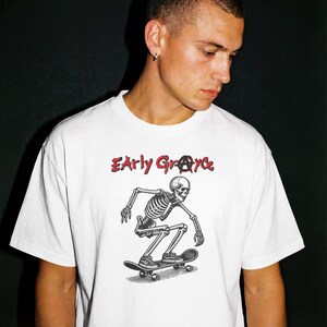 May include: White t-shirt featuring a black and white illustration of a skeleton skateboarding. The text "Early Grace" is printed in red above the skeleton. The shirt has a crew neck and short sleeves.