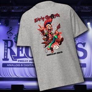 May include: A gray t-shirt featuring a cartoon punk rocker with a red mohawk and a guitar. The text "Early Grace" is above the graphic, and "american punk" is below. The shirt is displayed on a stage.