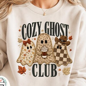 Könnte beinhalten: Ein cremefarbenes Sweatshirt mit dem Schriftzug COZY GHOST CLUB in schwarzen Buchstaben. Das Design zeigt drei Geister: einen mit Blumenmuster und Schleife, einen mit Leopardenmuster und Kaffeebecher sowie einen mit braunem Karomuster und Cowboyhut, umgeben von Herbstblättern.
