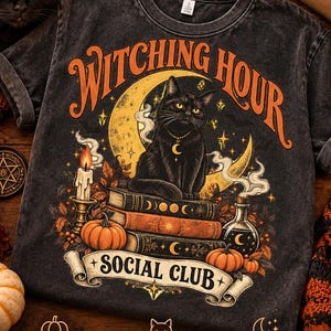 May include: A charcoal gray t-shirt featuring a graphic of a black cat sitting on a stack of books in front of a crescent moon. The text reads "WITCHING HOUR" and "SOCIAL CLUB". The design includes a candle, pumpkins, and smoke elements.