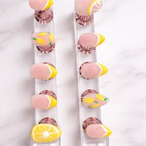 May include: A set of ten press-on nails arranged on two clear acrylic stands. The nails feature a nude base with various lemon-themed designs, including yellow French tips, hand-painted yellow lemon slices, and small green leaf accents.