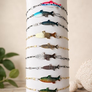 May include: A collection of fish-shaped bracelets in various colors and finishes, including pink, blue, gold, silver, and green. Each bracelet features a detailed fish pendant and a delicate chain. The bracelets are displayed on a white cylindrical stand.