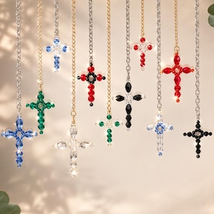 May include: A collection of beaded cross pendants in various colors, including blue, green, red, and black, hanging from silver and gold chains. The crosses are made of small, faceted beads, and are displayed against a soft, neutral background.