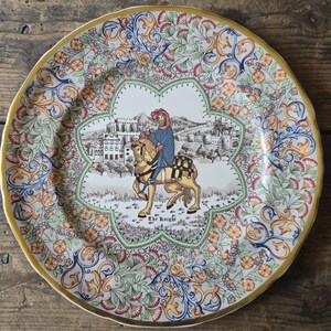 May include: A round ceramic plate featuring a central illustration of a knight on horseback with the text The Knight. The border is decorated with a colourful floral and scroll pattern in shades of blue, orange, red, and green with a gold rim.