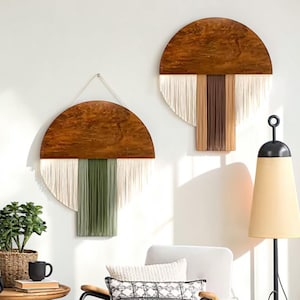 Puede incluir: Dos colgantes de pared semicirculares con una parte superior de madera y tela con flecos. Uno presenta flecos blancos y verdes, el otro blancos, marrones y beige. La decoración de la pared es de estilo bohemio moderno.