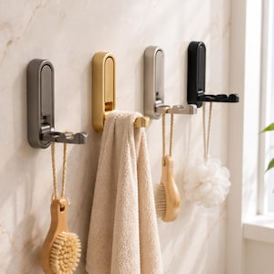 Op de afbeelding: Vier aan de muur gemonteerde haken in grijs, goud, zilver en zwart. Elke haak heeft een opvouwbare arm en houdt een ander item vast: een houten borstel, een handdoek, een andere borstel en een witte badspons. De haken zijn op een lichtgekleurde muur gemonteerd.
